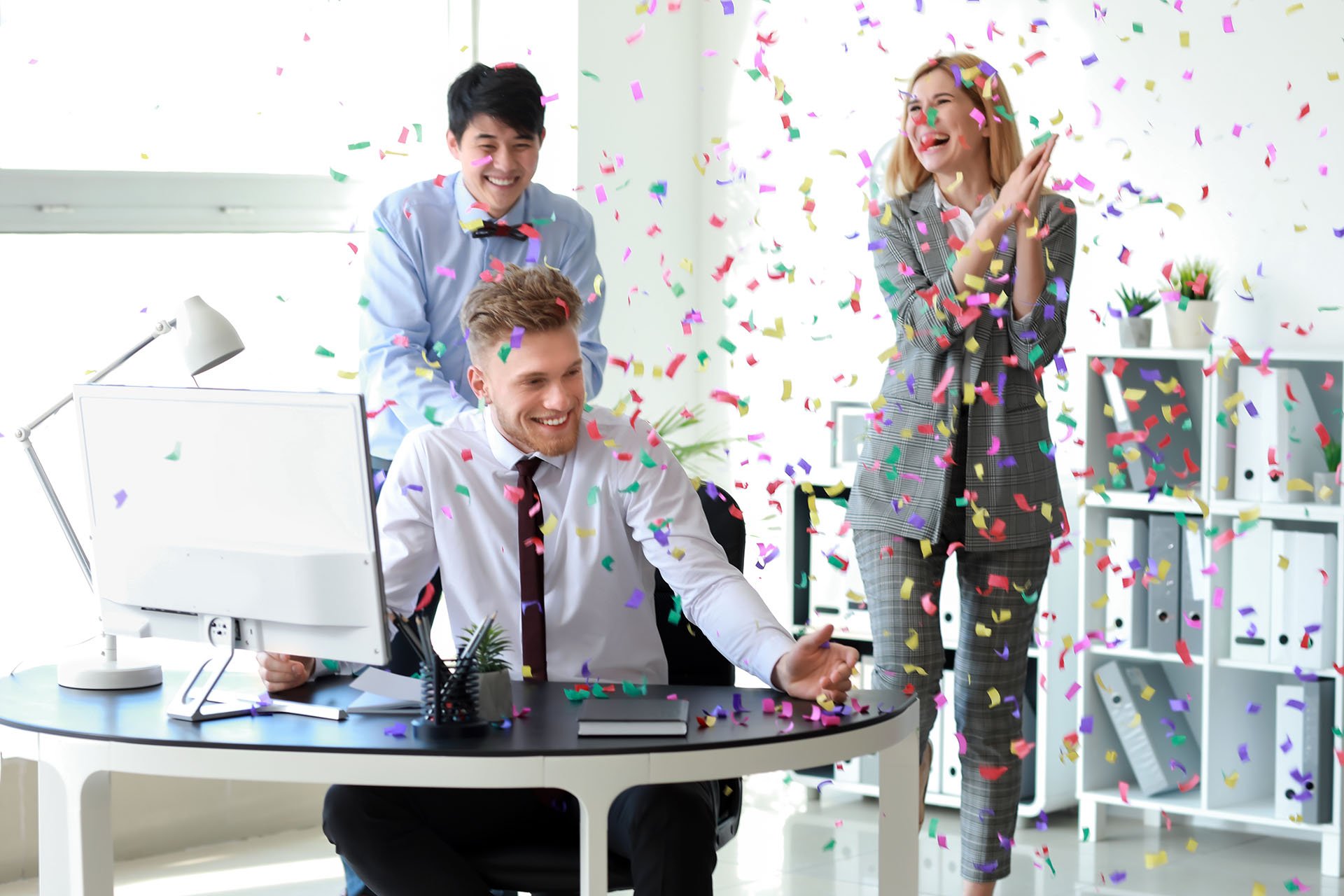 Two men and a woman are showered with confetti at a new employee's desk